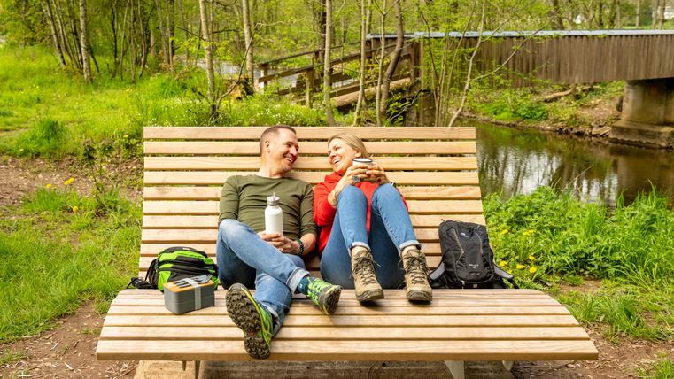 Ein Paar sitzt lächelnd auf einer Holzbank in der Natur. Im Hintergrund ist ein ruhiger Bach und eine Brücke zu sehen.
