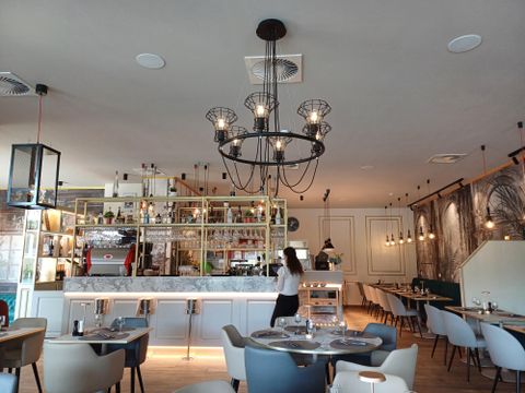 Een modern restaurant met een elegante inrichting en een stijlvolle kroonluchter. Aan de bar staan verschillende dranken, en de tafels zijn mooi gedekt.