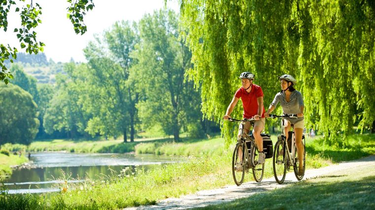 Two cyclists ride leisurely along a path, surrounded by lush greenery. In the background, a calm river flows.