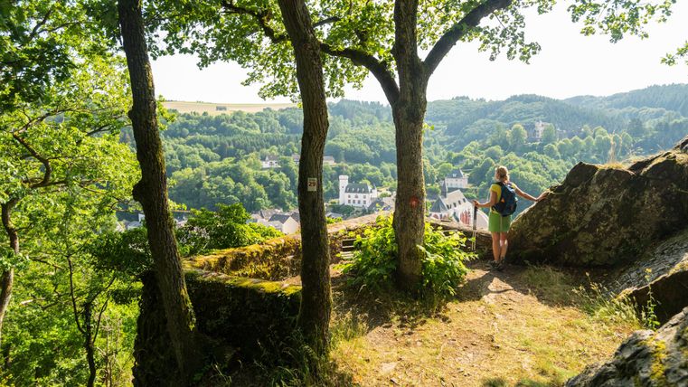 Une randonneuse se tient sur un rocher et regarde une vallée pittoresque avec des arbres verts et de petites maisons. Le soleil brille et le ciel est dégagé.