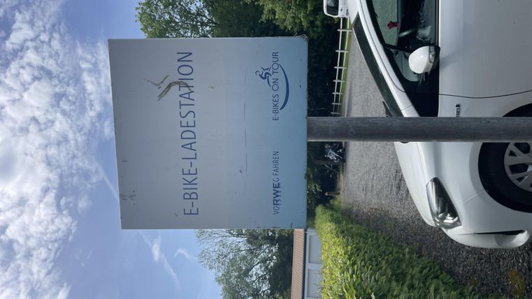 Een bord voor een E-bike oplaadstation staat op een rustige locatie. Op de achtergrond zijn bomen en een parkeerplaats zichtbaar.