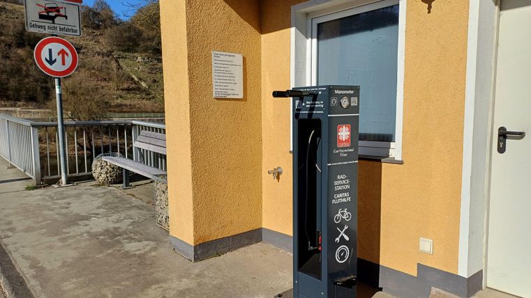 Eine Reparaturstation für Fahrräder steht an einer Gebäudewand. Im Hintergrund sind Verkehrsschilder und eine Brücke zu sehen.