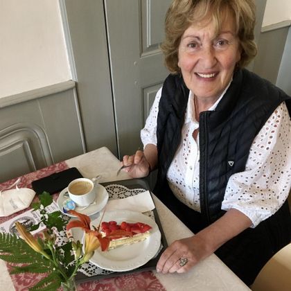 A friendly elderly woman is sitting at a table with a piece of cake and a cup of coffee. On the table, there is a small vase with a flower.