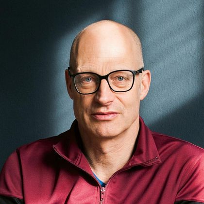 Een man met een bril en een glad hoofd in een donkerrood gekleurd bovenstuk. De achtergrond is in een zachtblauw met lichtschaduwen.