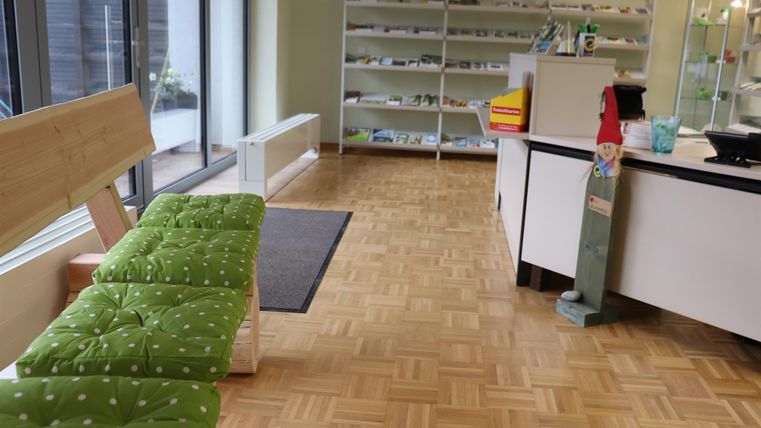 Une pièce lumineuse avec un sol en bois et de grandes fenêtres. Sur le côté gauche se trouve un banc vert et un comptoir d'accueil sur le côté droit.