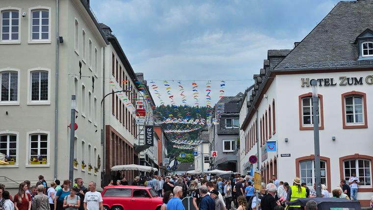 Une rue animée avec de nombreuses personnes et des banderoles colorées. Des bâtiments historiques se tiennent de part et d'autre, et des tables de cafés sont installées.