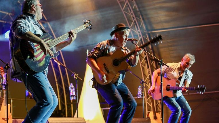 Trois musiciens jouent de la guitare acoustique sur scène. L'atmosphère est vivante et festive avec des lumières colorées en arrière-plan.
