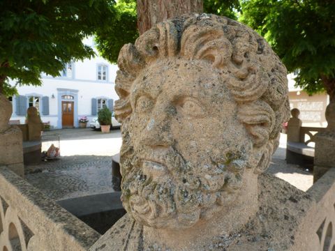 Une antique bustes en pierre avec une représentation détaillée d'un homme barbu. En arrière-plan, on aperçoit des arbres verts et un bâtiment historique.