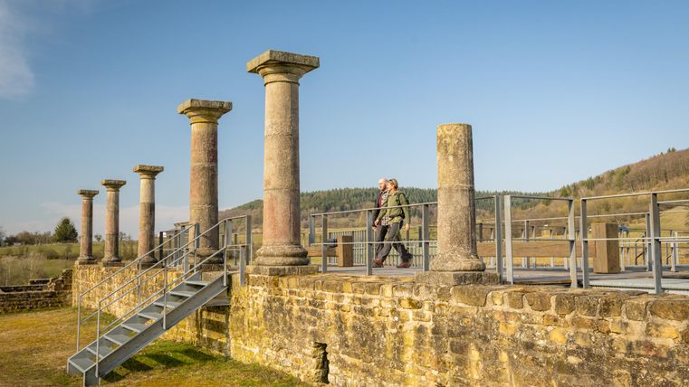 Eine historische Ruine mit Säulen und einer Aussichtsplattform. Im Hintergrund sind sanfte Hügel und ein klarer Himmel sichtbar.