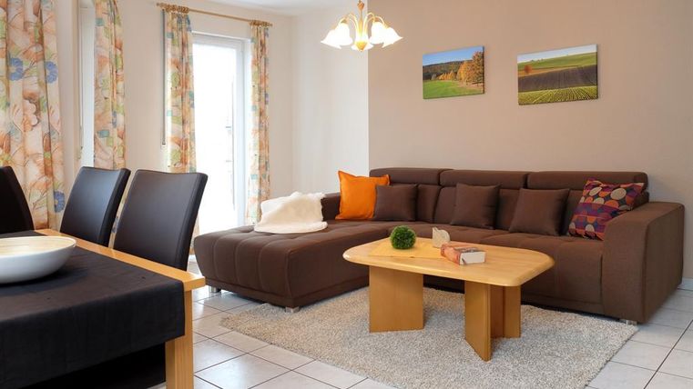 A bright living room with a cozy sofa and a wooden table. Pictures hang on the walls, and the windows are decorated with colorful curtains.