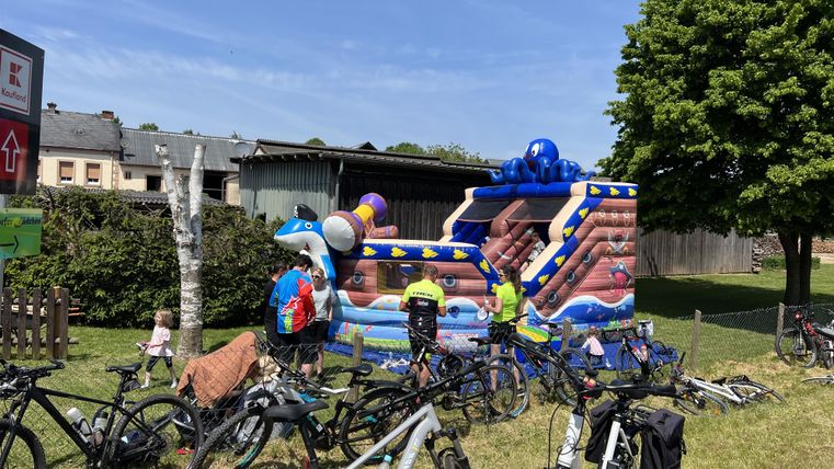 Een kleurrijk springkasteel staat in een groene tuin, omringd door fietsen. Kinderen hebben plezier met spelen.
