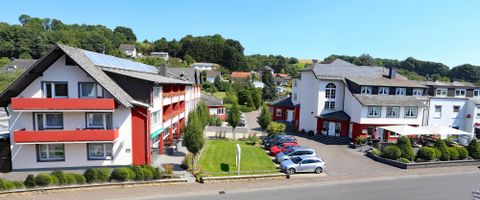 A modern hotel building with red accents and a clear sky. The surroundings are green and inviting, with cars in the parking lot.