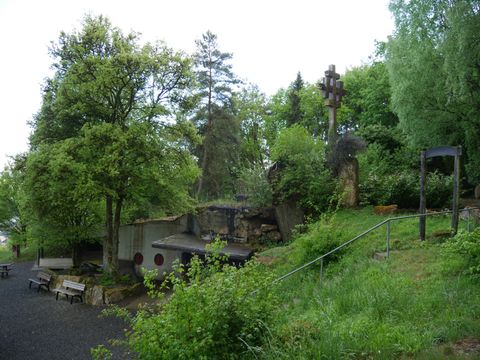 Un paysage pittoresque avec de nombreux arbres et un environnement verdoyant. Au premier plan, des bancs et quelques sculptures en pierre sont visibles.