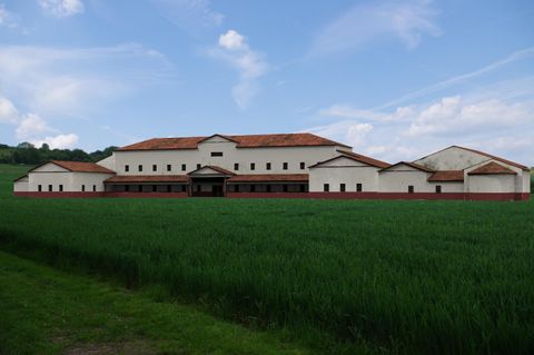 Un grand bâtiment moderne avec un toit rouge devant un champ vert. Le ciel est bleu avec quelques nuages.