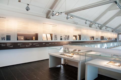 Ein modernes Museum mit einer Wand voller Informationen und Exponaten. Im Vordergrund sind Vitrinen mit verschiedenen Objekten zu sehen.