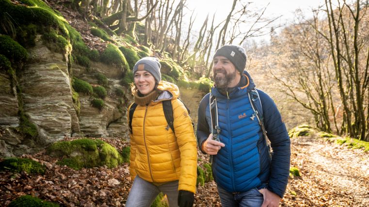 Two hikers on a beautiful forest path with green moss and rocks. It seems to be a sunny day and the nature around them is vibrant.