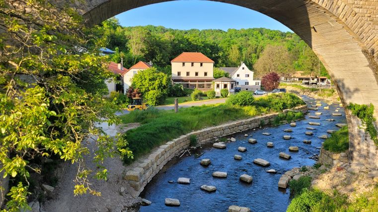 A picturesque river flows under a stone bridge. In the background, some cozy houses and green riverside land can be seen.
