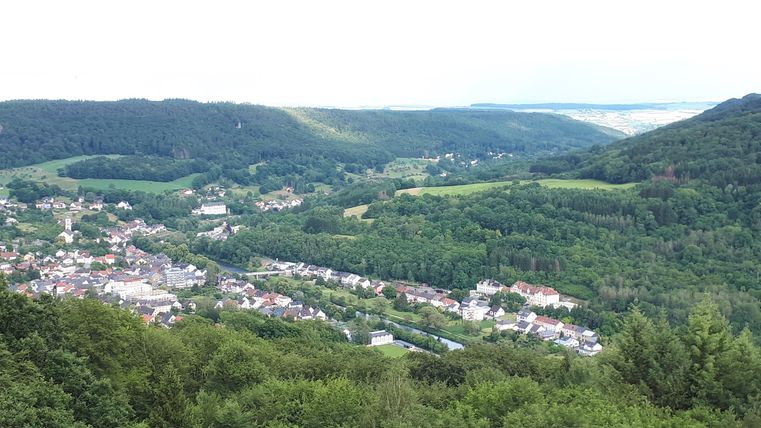 Eine malerische Aussicht auf ein Dorf, umgeben von grünen Hügeln und Wälder. Der Himmel ist bewölkt, aber die Landschaft wirkt friedlich.