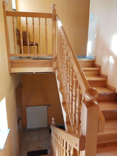 A bright staircase with a wooden railing leads upwards. In the background, a chair and a wall in warm colors can be seen.