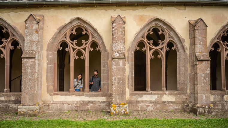 Two people are standing in an old building with beautiful Gothic windows. The surroundings are green and well-kept.