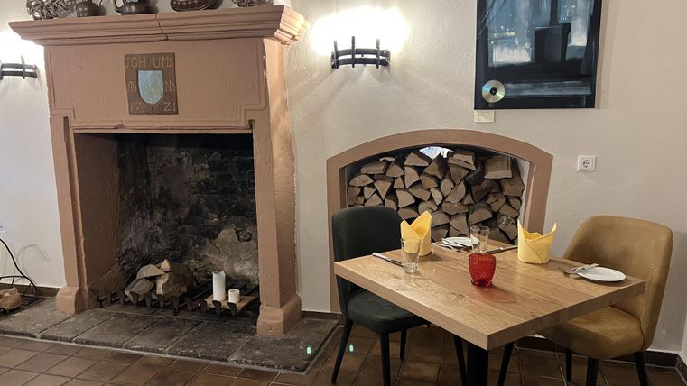 Een gezellige restaurantruimte met een haard en een houtopslag. Op de tafel liggen gele servetten en een rood glas.