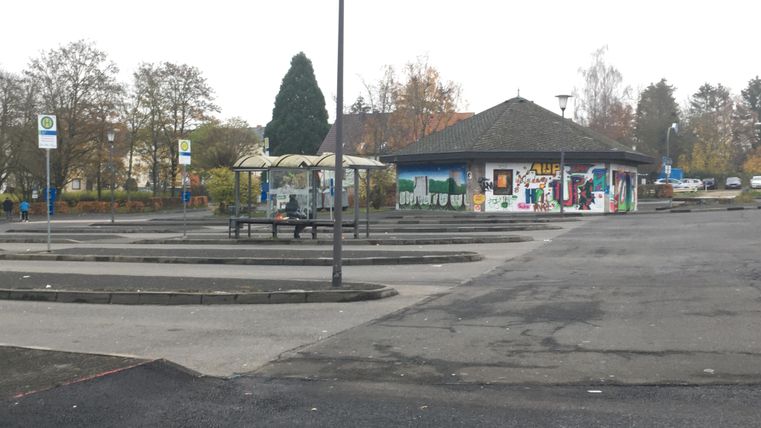 Ein leerer Parkplatz mit einer kleinen, bunten Gebäudefassade im Hintergrund. Es ist bewölkt, und einige Bäume sind in der Nähe sichtbar.