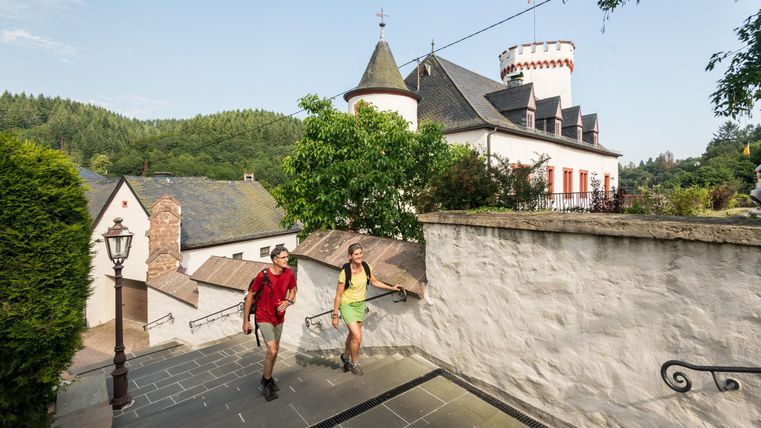Two hikers are going down a staircase, surrounded by beautiful scenery. In the background, there is a charming castle with a onion tower and red windows.