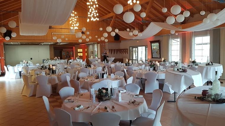 Ein festlich dekorierter Raum mit runden Tischen und weißer Tischdecke. Die Decke ist mit leuchtenden Kugeln und Stoffbahnen geschmückt.