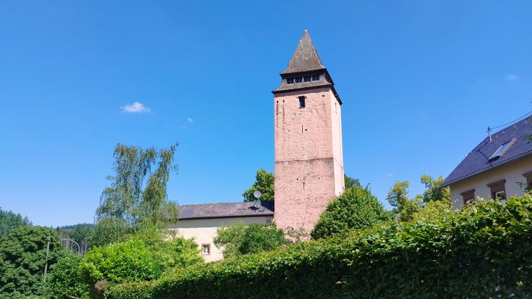 Ein historischer Turm aus rotem Stein unter klarem, blauem Himmel. Umgeben von Grünpflanzen und Bäumen.