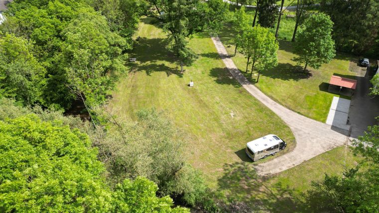 Een groene weide omringd door bomen, met een klein pad dat door het landschap leidt. Op de achtergrond staat een camper en een picknicktafel.