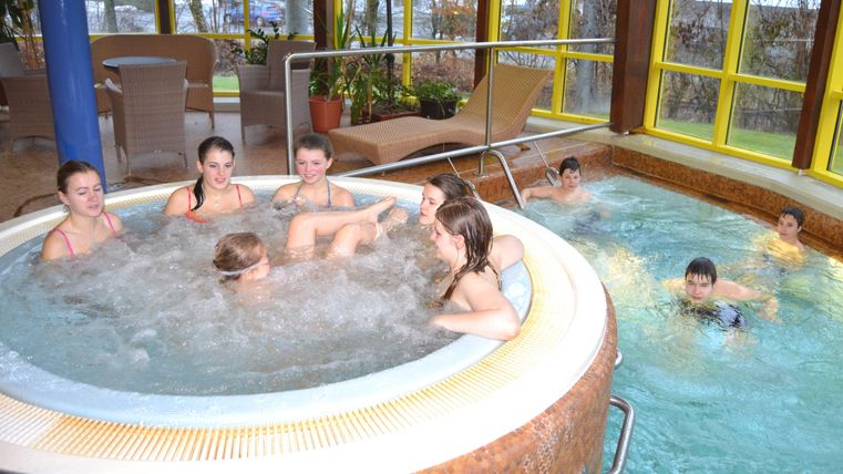 A group of children and teenagers relaxes in the hot tub. In the background, other children are swimming in the pool.