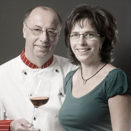 A chef in a white chef's jacket holds a wine glass and stands next to a smiling woman. Both are looking at the camera and appear friendly and inviting.
