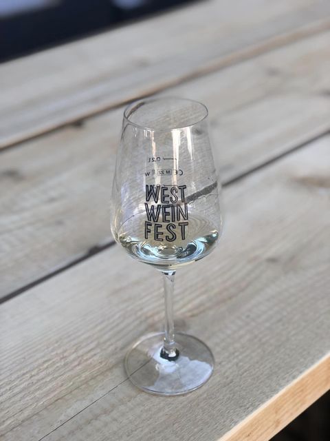 Een glas met de opdruk "WEST WEINFEST" staat op een houten tafel. De tafel heeft een natuurlijke houtoppervlak en creëert een gezellige sfeer.