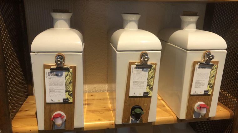 Trois conteneurs blancs sur une étagère, remplis d'huile ou de vinaigre. Chaque conteneur a une étiquette et un bec verseur.