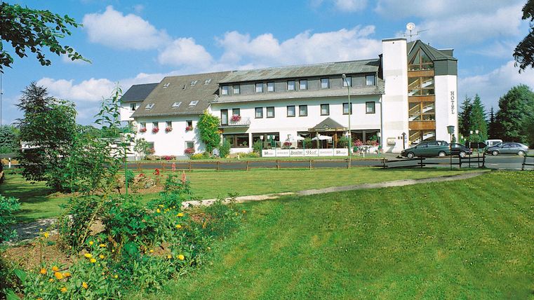 A modern hotel building surrounded by a well-maintained lawn and green plants. The sky is blue with some clouds.