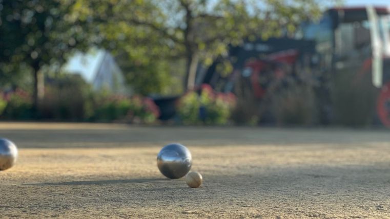 Drie metalen ballen liggen op een zandplek. Op de achtergrond zijn groene bomen en een rode tractor te zien.