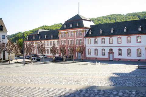 A historic building with red and white facades. The square in front is paved with stones and lined with trees.