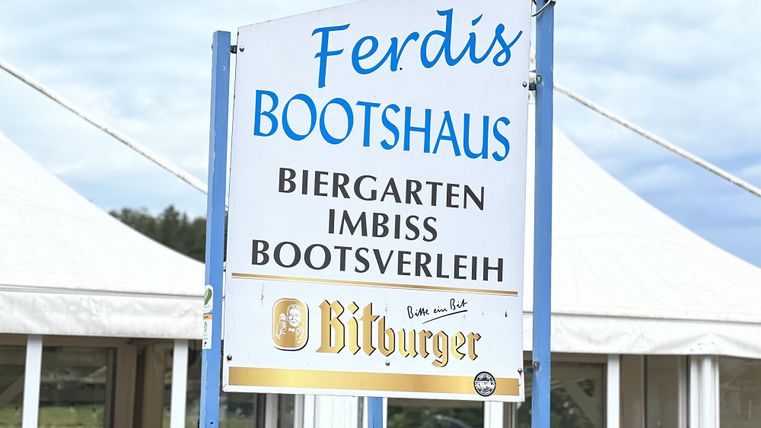 Een bord voor de Ferdi's Bootshaus, dat een biergarten, een snackbar en bootverhuur aanbiedt. De omgeving is uitnodigend en vriendelijk.