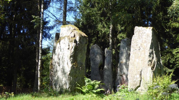 A group of large stones stands in a green meadow, surrounded by trees. The sun shines through the leaves, creating a peaceful atmosphere.