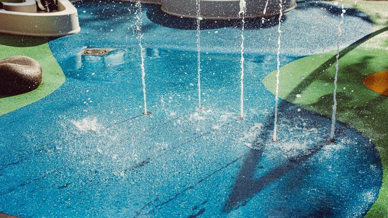 Een kleurrijke waterspeelplaats met spuitende waterfonteinen. De bodem is in verschillende kleuren vormgegeven en geeft een vrolijke sfeer.