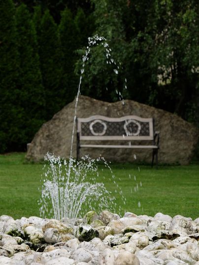 A beautiful garden with a fountain of water and a bench in the background. The surroundings are green and inviting.