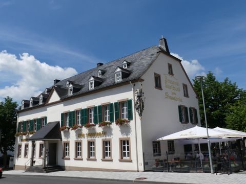 A lovely two-story building with green shutters and flower boxes. In front of the house, there is a terrace with umbrellas and an inviting seating area.