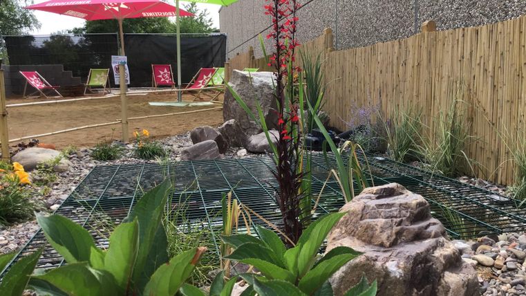 Een tuingebied met stenen en planten, omringd door een houten hek. Twee zonneschermen in groen en rood bieden schaduw en een ontspannen sfeer.