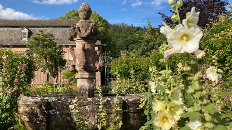 Ein idyllischer Garten mit einem alten Brunnen und schönen Blumen. Im Hintergrund ist ein historisches Gebäude und ein blauer Himmel zu sehen.