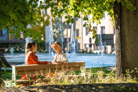 Zwei Frauen sitzen auf einer Bank im Park und unterhalten sich. Im Hintergrund sind ein Brunnen und grüne Bäume zu sehen.