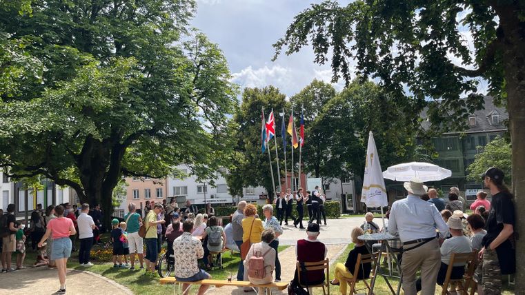 A group of people gathers in a park to watch a performance. Colorful flags are set up, and there is a festive atmosphere.