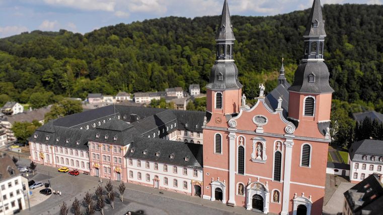 Une église impressionnante avec deux hautes tours se dresse au centre, entourée de bâtiments historiques et d'une grande place. À l'arrière-plan, des collines verdoyantes sont visibles et le ciel est partiellement nuageux.