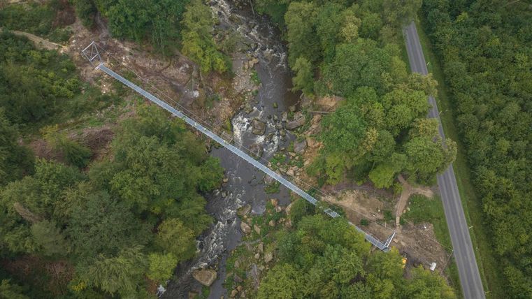 Eine Brücke über einen Fluss, umgeben von dichtem Wald. Im Hintergrund ist eine Straße sichtbar.