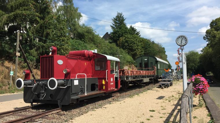 Een rode locomotief staat naast een ouder wagon op een spoor. Op de achtergrond zijn bomen en een kloktoren te zien.