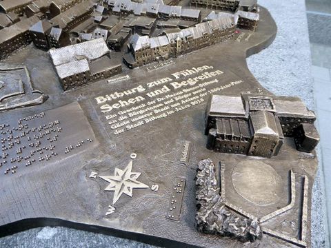 Eine detailreiche Bronzeansicht einer Stadt mit Gebäuden und Straßen. Der Text hebt die Bedeutung der Stadt hervor und lädt ein, sie zu erkunden.